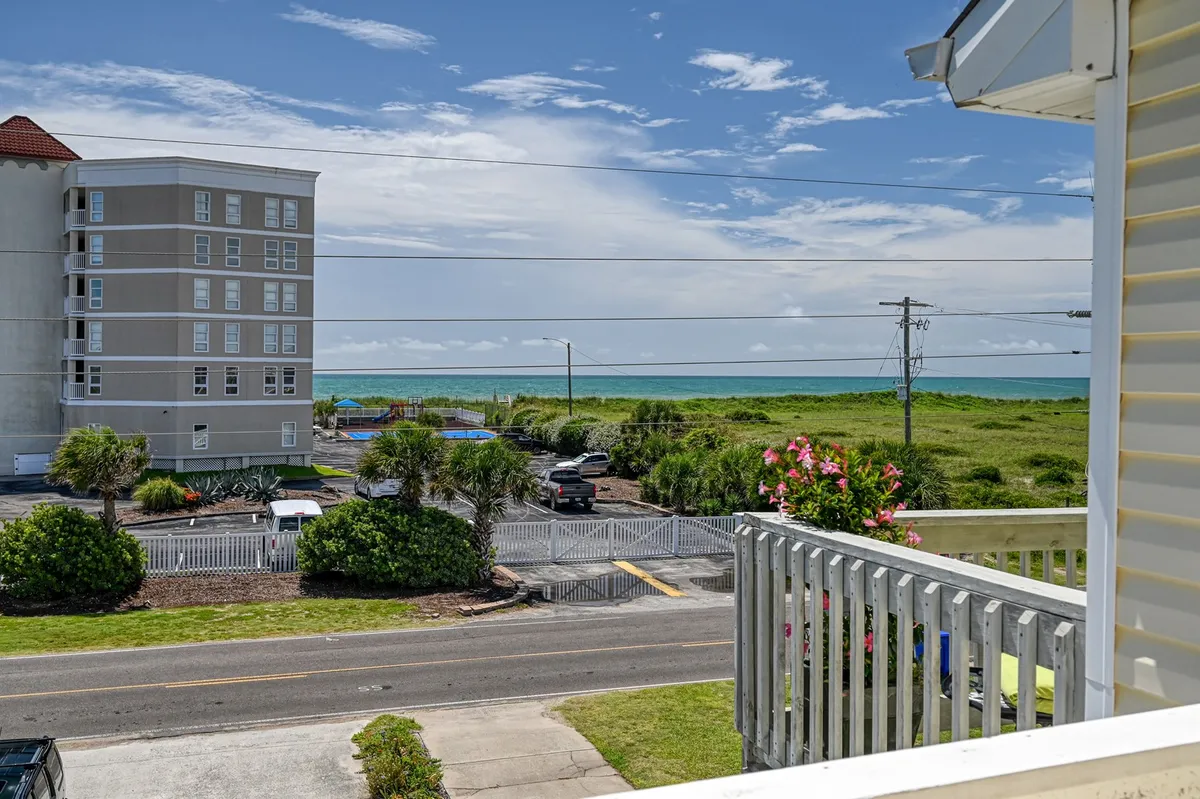 The image showcases a charming single-family house with a welcoming facade, surrounded by lush flowering plants and greenery. The home's balcony faces the sparkling Atlantic Ocean, offering breathtaking sunrise views. The scene also features a neatly-kept parking area, with the ocean just steps away—perfect for easy beach access. The mood is tranquil, inviting viewers to imagine relaxing mornings on the balcony or taking peaceful walks along the coast.