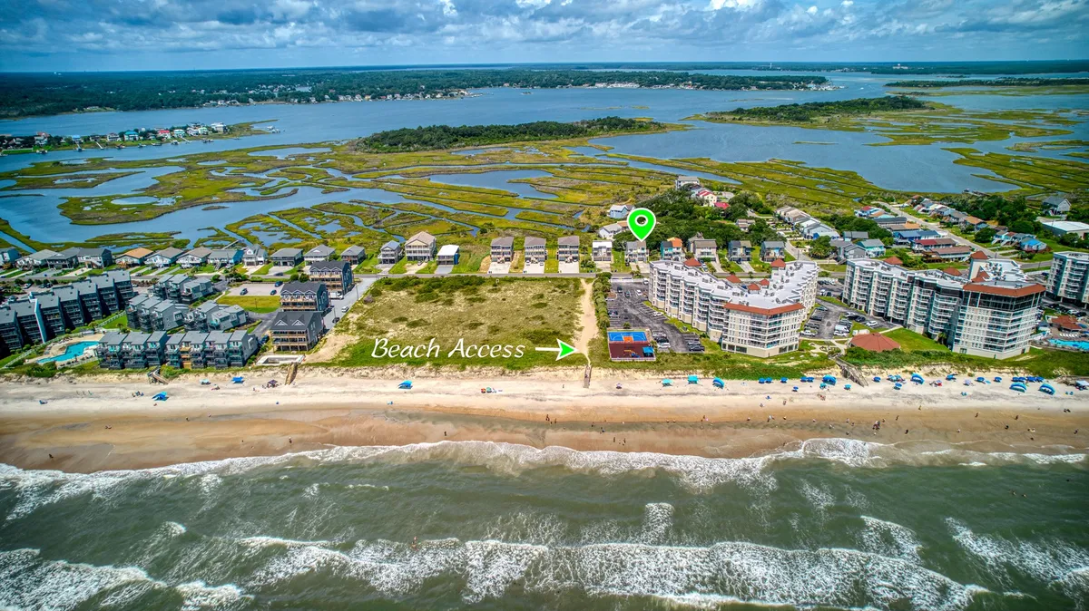 This exterior image showcases direct beach access with a clear view of the sandy path leading to the ocean, gentle waves in the background, and coastal buildings nearby. Marshland hints at the surrounding natural beauty, emphasizing the home's proximity to outdoor adventure.
