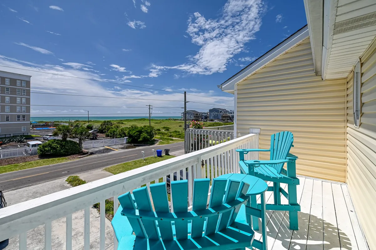 The scene features a pair of turquoise Adirondack chairs on a deck or patio with an expansive view of open landscape and blue sky dotted with clouds. This tranquil, elevated vantage point suggests a peaceful spot for watching sunrise, sunset, or simply relaxing after a beach outing.