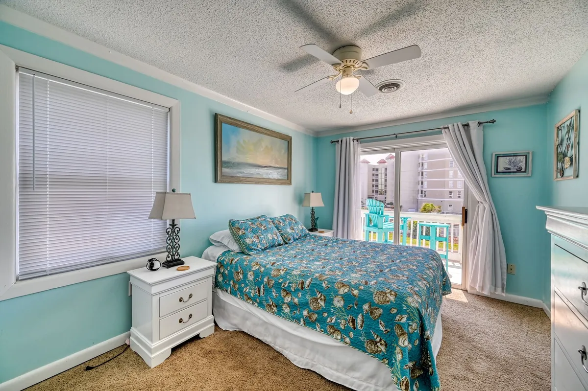 This image features a serene bedroom with a bed dressed in patterned bedding, a large window offering access to a balcony, and artwork on the wall. The atmosphere is peaceful and inviting, with clear oceanview indications.