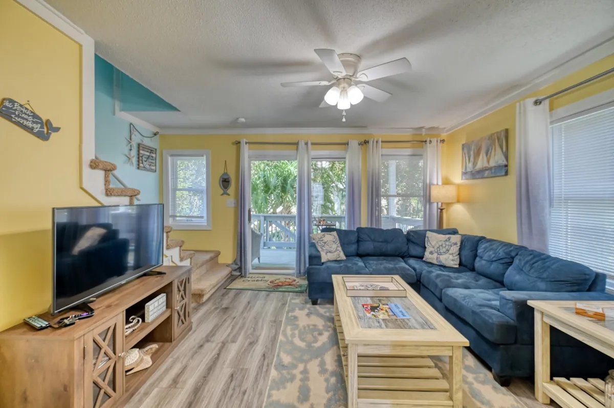A sunny, welcoming living room featuring a blue sectional sofa arranged around a wooden coffee table, with a large television as the focal point. Expansive windows let in abundant natural light and offer a lush green view, creating an inviting space for relaxing or socializing.