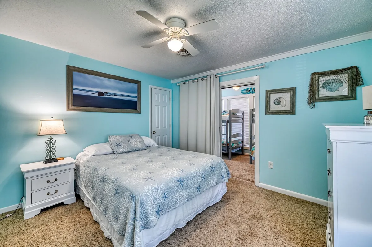This interior image showcases a bed with a patterned comforter, a lamp on the bedside table, ocean-themed artwork, and, crucially, bunk beds visible through a curtain. The room is bathed in natural light and exudes a cozy, family-friendly atmosphere.