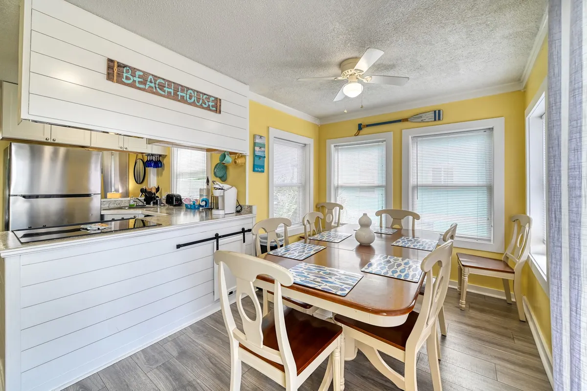 A cheerful open-concept dining and kitchen area, featuring a traditional dining table for eight, a spacious kitchen counter, large windows, and a prominent 'Beach House' sign. The space feels lively and perfect for gatherings, family meals, or entertaining.