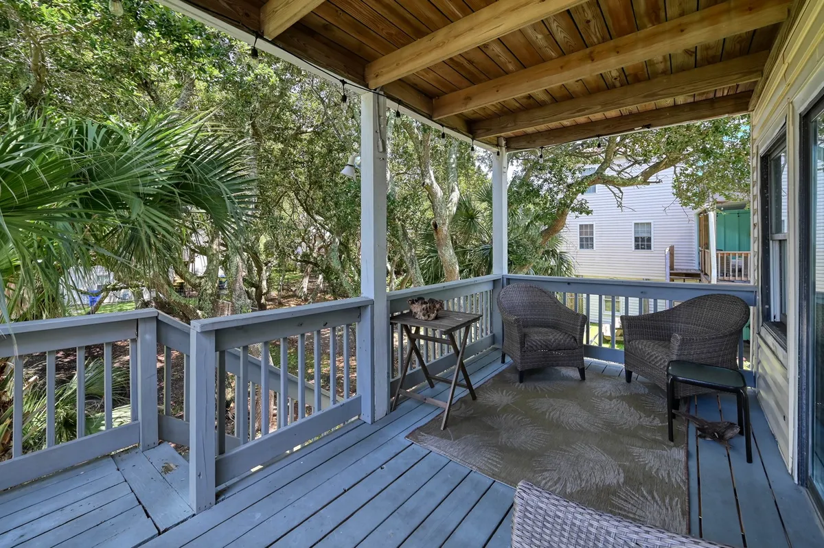 A tranquil backyard scene featuring all-weather wicker chairs and a small table arranged beneath the shade of dense greenery, including palms and bay bushes. Decorative touches on the table and the inviting setup suggest a comfortable outdoor lounge space perfect for both people and pets.